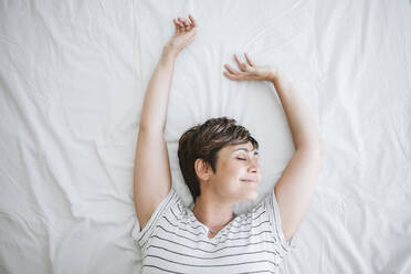 Smiling woman with arms raised lying on bed at home - EBBF04647