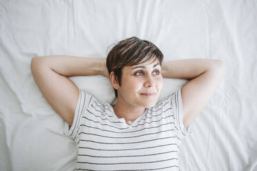 Thoughtful woman with hands behind head lying on bed at home - EBBF04649