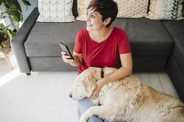 Woman holding smart phone while sitting with Golden Retriever at home - EBBF04652