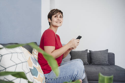 Mid adult woman holding smartphone while sitting on sofa in living room - EBBF04665