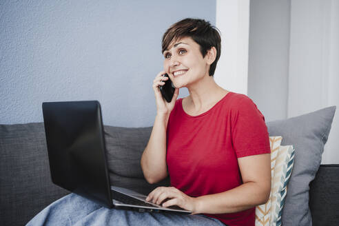 Aufgeregte Geschäftsfrau, die mit ihrem Smartphone spricht, während sie zu Hause am Laptop sitzt - EBBF04669