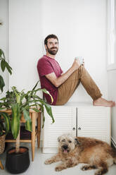 Mid adult man looking away while sitting with mixed-breed dog at home - EBBF04682
