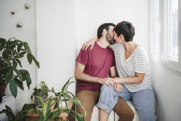 Mann und Frau küssen sich, während sie in der Nähe von Pflanzen zu Hause sitzen - EBBF04683