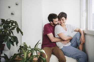 Mid adult man embracing woman while sitting near window at home - EBBF04684