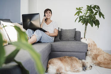 Geschäftsfrau mit Haustieren, die mit ihrem Smartphone telefoniert, während sie zu Hause auf dem Sofa mit ihrem Laptop sitzt - EBBF04695