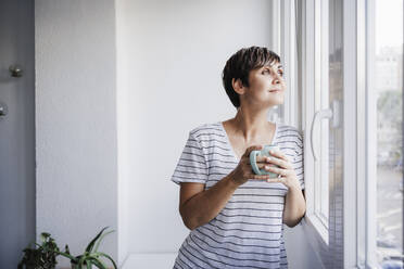 Woman holding mug while looking through window at home - EBBF04699