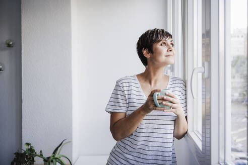 Frau hält Tasse, während sie durch ein Fenster zu Hause schaut - EBBF04699