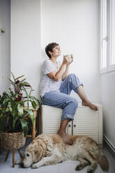 Woman holding mug while sitting by dog at home - EBBF04703