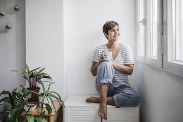 Smiling mid adult woman with mug looking through window while sitting at home - EBBF04710