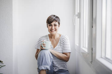 Glückliche Frau hält Tasse, während sie zu Hause am Fenster sitzt - EBBF04711