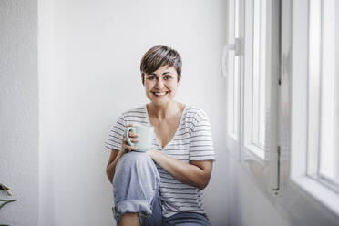 Happy woman holding mug while sitting near window at home - EBBF04711