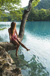 Junge Frau sitzt auf einem Baum, der am Seeufer im Nationalpark Plitvicer Seen wächst - JAQF00823