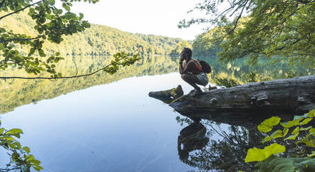 Female hiker crouching on fallen lakeshore tree in Plitvice Lakes National Park - JAQF00836