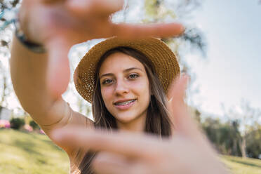 Lächelnder weiblicher Teenager mit Zahnspange, der die Geste des Fotografierens demonstriert, während er in die Kamera schaut, tagsüber auf unscharfem Hintergrund - ADSF30552