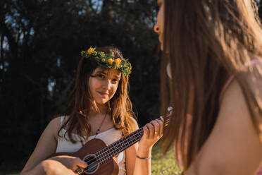 Teen in flower wreath playing ukulele while sitting against best female friend with wireless headphones in back lit - ADSF30555