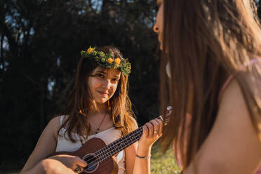 Teenager mit Blumenkranz spielt Ukulele, während er gegen seine beste Freundin mit drahtlosen Kopfhörern in der Rückenlehne sitzt - ADSF30555