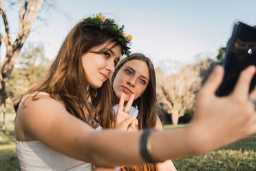 Weibliche Teenager demonstrieren die Geste des Sieges, während sie ein Selbstporträt mit dem Handy in einem sonnigen Park mit unscharfem Hintergrund aufnehmen - ADSF30557