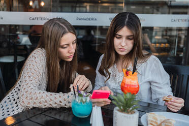 Beste Freundinnen mit Gläsern erfrischender Getränke, die am Tisch in einem städtischen Café auf ihrem Handy surfen - ADSF30564