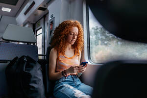 Interested woman with curly hair in ripped jeans text messaging on cellphone during trip on train in daytime - ADSF30566