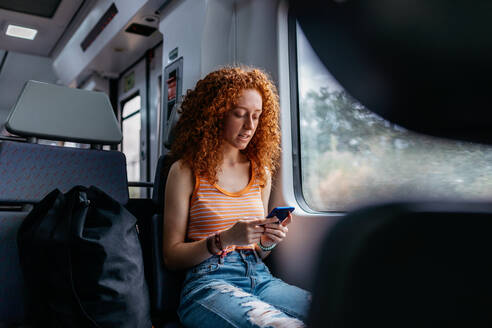 Interessierte Frau mit lockigem Haar in zerrissenen Jeans, die während einer Zugfahrt tagsüber eine Textnachricht auf ihrem Mobiltelefon sendet - ADSF30566