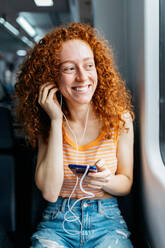 Candid young woman with curly red hair and cellphone listening to song from earphones while looking away on train - ADSF30567