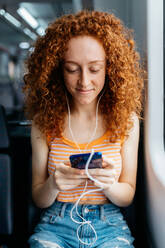 Interested woman with curly hair in ripped jeans text messaging on cellphone during trip on train in daytime - ADSF30568