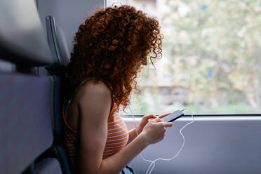 Interessierte Frau mit lockigem Haar in zerrissenen Jeans, die während einer Zugfahrt tagsüber eine Textnachricht auf ihrem Mobiltelefon sendet - ADSF30570