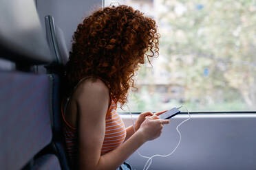 Interested woman with curly hair in ripped jeans text messaging on cellphone during trip on train in daytime - ADSF30570