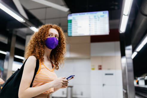 Side view of woman in textile mask with cellphone and rucksack looking away on subway platform - ADSF30572