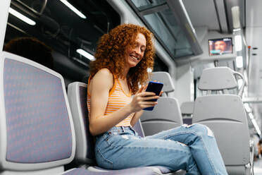 Interested woman with curly hair in ripped jeans text messaging on cellphone during trip on train in daytime - ADSF30576