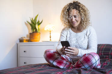 Lächelnde reife Frau, die ein Smartphone benutzt, während sie im Schneidersitz auf dem Bett sitzt - SIPF02465