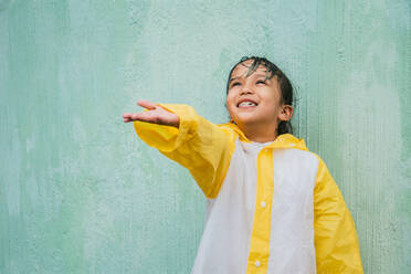 Charmante ethnischen Kind in slicker mit Palme oben fangen regen, während der Blick nach oben auf Pastell Hintergrund - ADSF30611