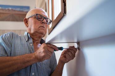 Attentive elderly male in eyeglasses with manual screwdriver screwing shelf to wall in house room - ADSF30616