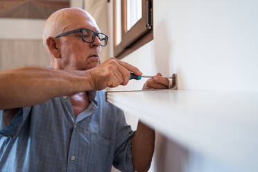 Aufmerksamer älterer Mann mit Brille und Schraubenzieher schraubt ein Regal an die Wand eines Wohnzimmers - ADSF30617