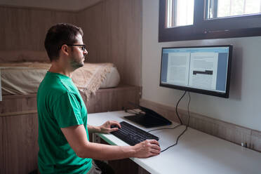 Seitenansicht eines männlichen Bloggers mit Brille, der Text auf einem Monitor bearbeitet, während er auf einer Tastatur in einem Hauszimmer tippt - ADSF30620