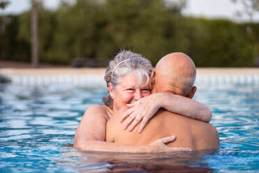 Lächelnde grauhaarige Frau umarmt kahlköpfigen Mann beim Schwimmen in sauberem Poolwasser zusammen - ADSF30622
