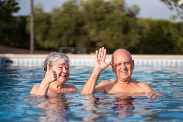 Cheerful senior couple waving while swimming in pool and looking at camera - ADSF30623