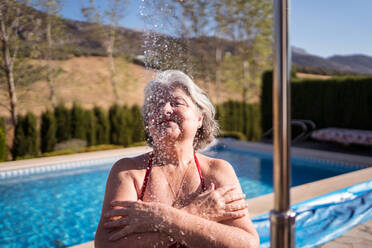 Fröhliche ältere Frau im Bikini genießt die Spritzer aus der Dusche in der Nähe des Pools mit transparentem klarem Wasser - ADSF30624
