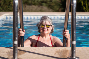 Positive ältere Frau in Badekleidung und Sonnenbrille, die im Schwimmbad abtaucht und sich an den Edelstahlhandläufen festhält, während sie sich an einem sonnigen Tag entspannt - ADSF30631