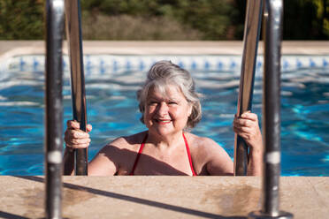 Positive ältere Frau in Badekleidung, die im Schwimmbad nach unten geht und sich an den Handläufen aus Edelstahl festhält, während sie sich an einem sonnigen Tag entspannt - ADSF30632