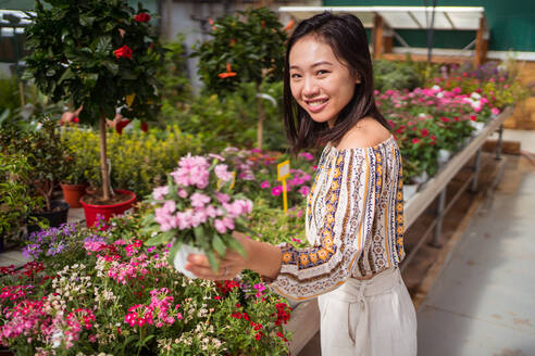 Aufrichtige junge ethnische weibliche Käuferin wählt blühende Blumen mit angenehmem Duft in einem Gartengeschäft am Tag aus - ADSF30645