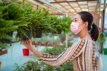 Side view of young ethnic female buyer in sterile mask choosing potted plant in garden center - ADSF30646