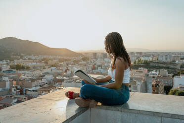 Trendy ethnic female in sandals reading textbook while sitting with crossed legs on fence in town - ADSF30658