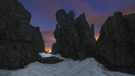 Panoramablick auf Tourist mit Fackel auf sandigem Land zwischen rauen Bergen unter bewölktem Himmel mit Sternen bei Sonnenuntergang - ADSF30660