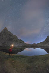 Rückenansicht eines nicht erkennbaren männlichen Touristen mit Taschenlampe, der verschneite Berge bewundert, die sich im Wasser unter dem nächtlichen Sternenhimmel spiegeln - ADSF30664