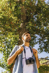 Junger Mann mit Kamera unter einem Baum an einem sonnigen Tag - MGRF00504