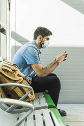 Man using mobile phone while sitting on bench during COVID-19 - MGRF00505