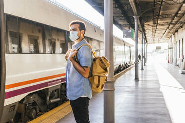 Junger Mann mit Rucksack auf dem Bahnsteig während der Pandemie - MGRF00506