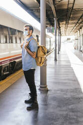 Young man wearing protective face mask standing on railroad station platform - MGRF00507