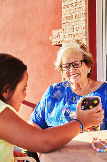 Glückliche Enkelin sitzt am Tisch und zeigt der lächelnden Großmutter Fotos auf dem Smartphone, während sie auf der Terrasse in der Sonne sitzt - ADSF30685